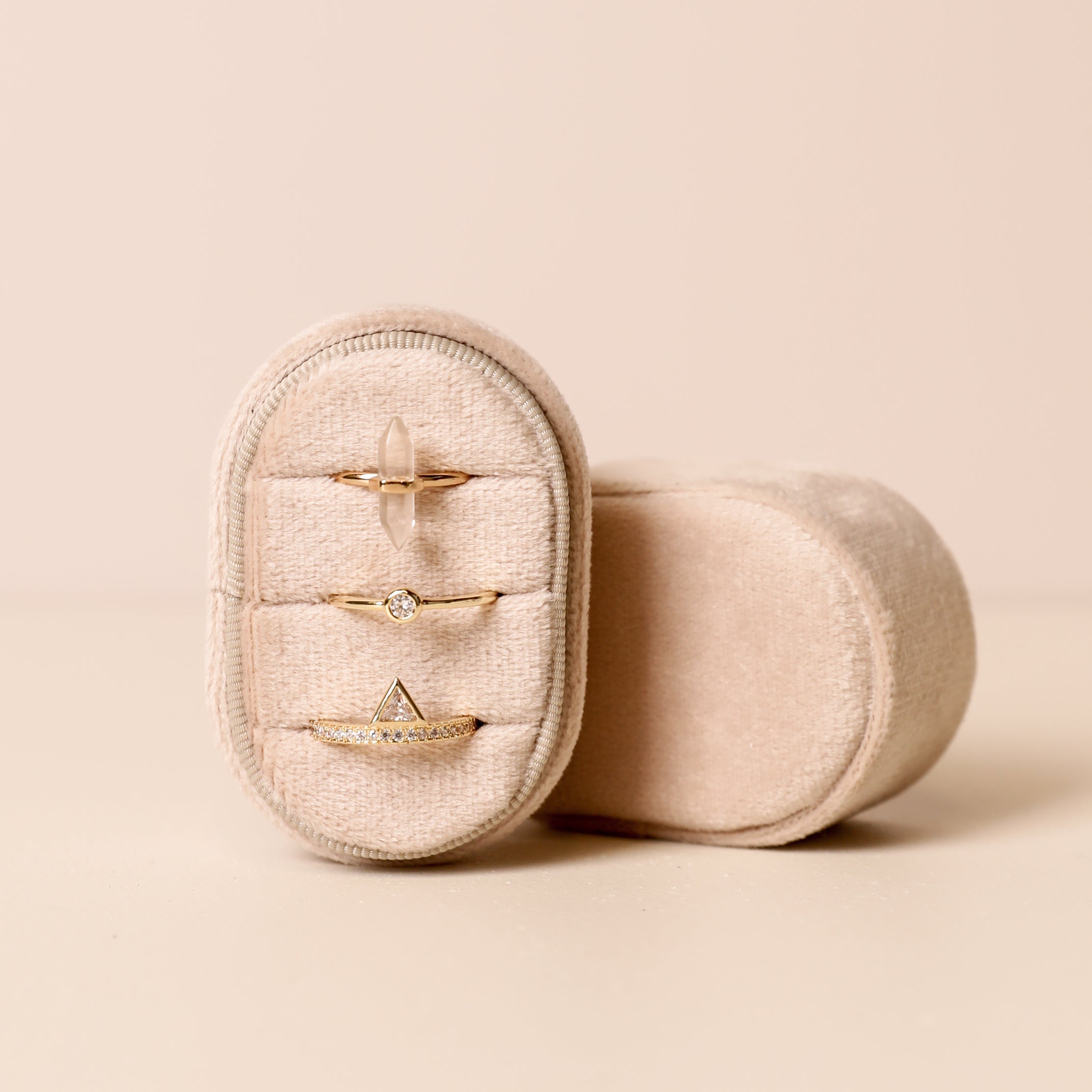 The Small Oval Velvet Jewelry Box holds three delicate gold rings with geometric designs, displayed against a soft beige background.
