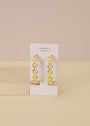 The Daisy Chain Earrings - Enamel and Gold are showcased on a white JaxKelly card, featuring 18K gold plating and delicate flower designs, set against a soft beige background.