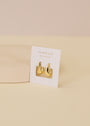 A pair of Small Ripple Gold Hoop earrings with 18K gold plating, featuring an abstract shape, are displayed on a white JAXKELLY WEAR GOOD VIBES card atop a beige surface against a neutral background.