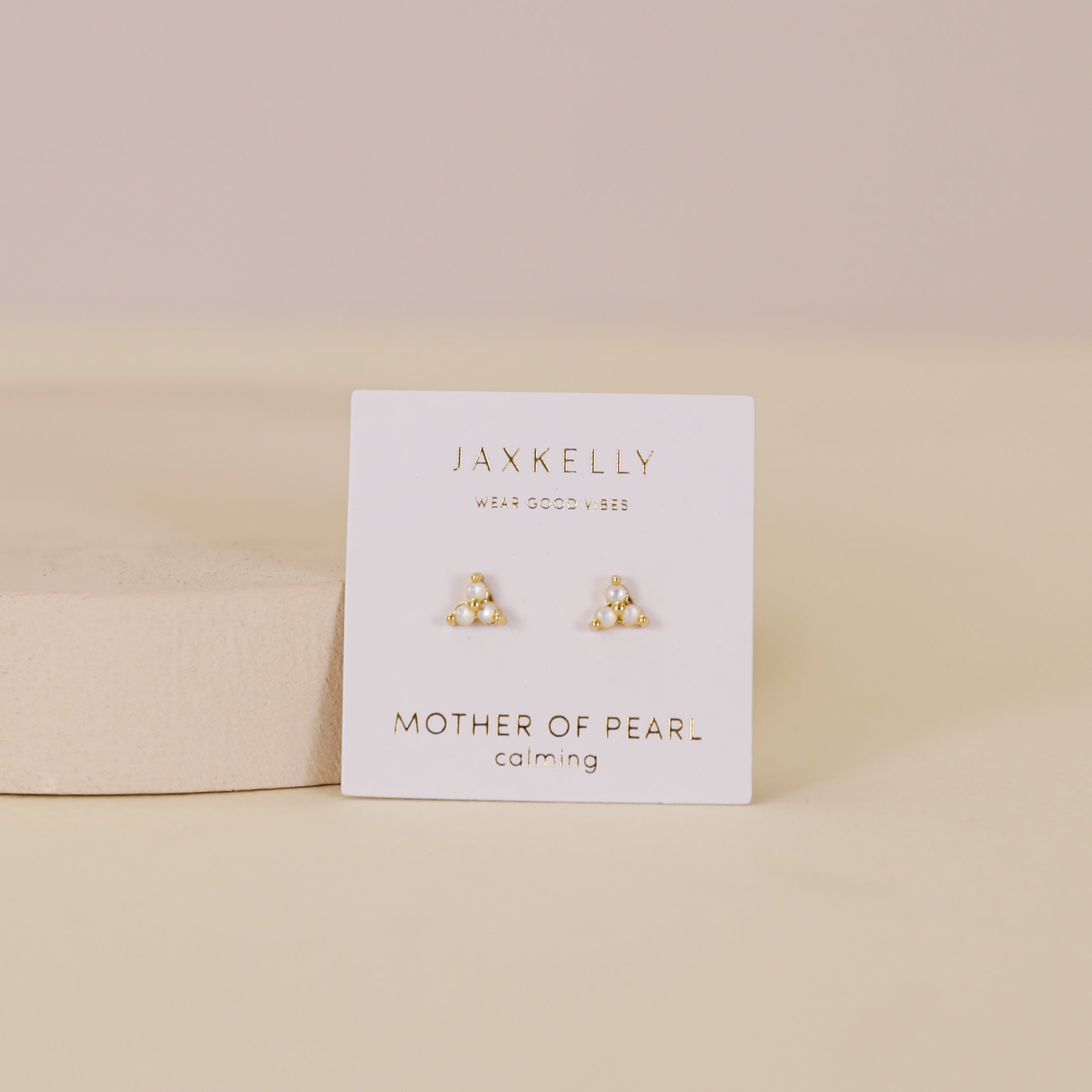 The Trio Cluster Gold Earrings - Pearl by JaxKelly are displayed on a white card reading "Mother of Pearl, calming," set against a soft beige background.