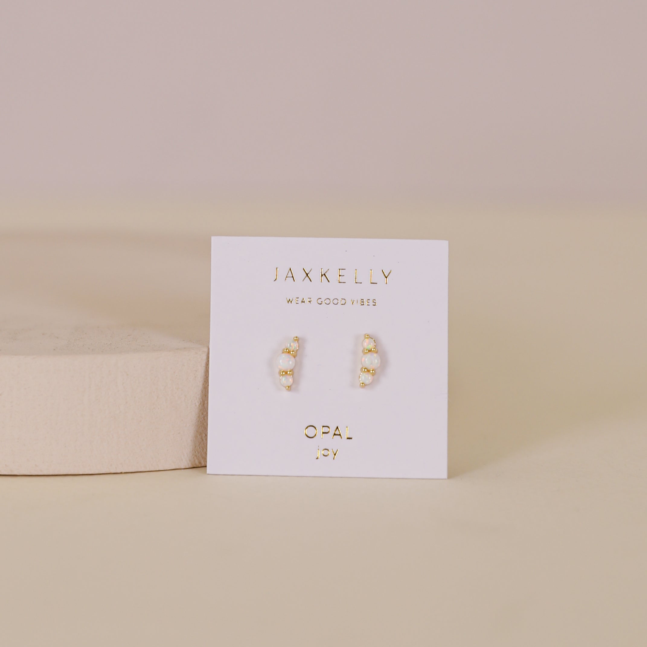 A pair of Crescent Gold Earrings - White Opal, featuring small gold-plated studs with lab-grown opals, is displayed on a white card labeled JaxKelly Opal against a soft beige background.