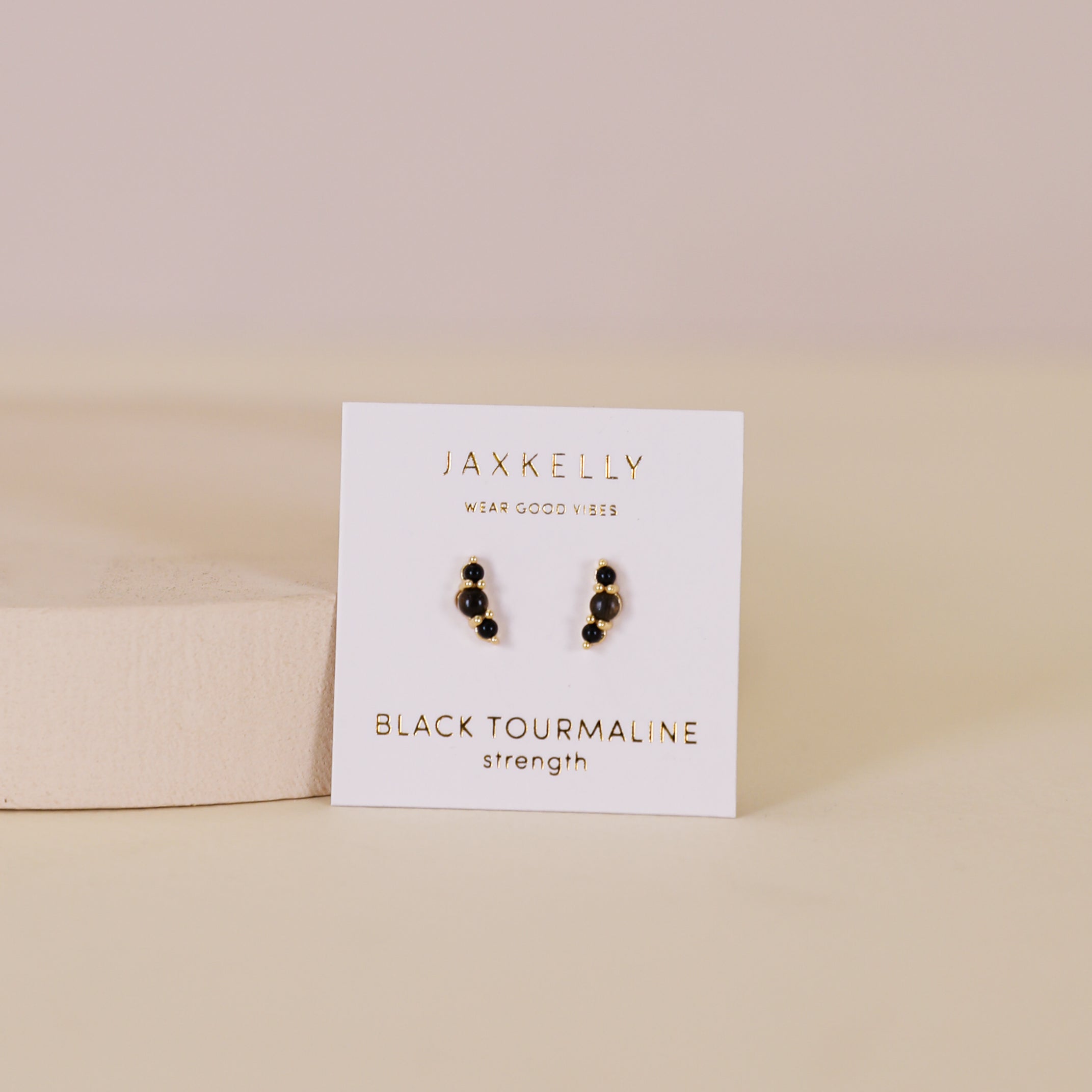A pair of Crescent Gold Earrings - Black Tourmaline is displayed on a white JaxKelly card labeled "Black Tourmaline Strength," set against a light beige surface and a soft beige background.