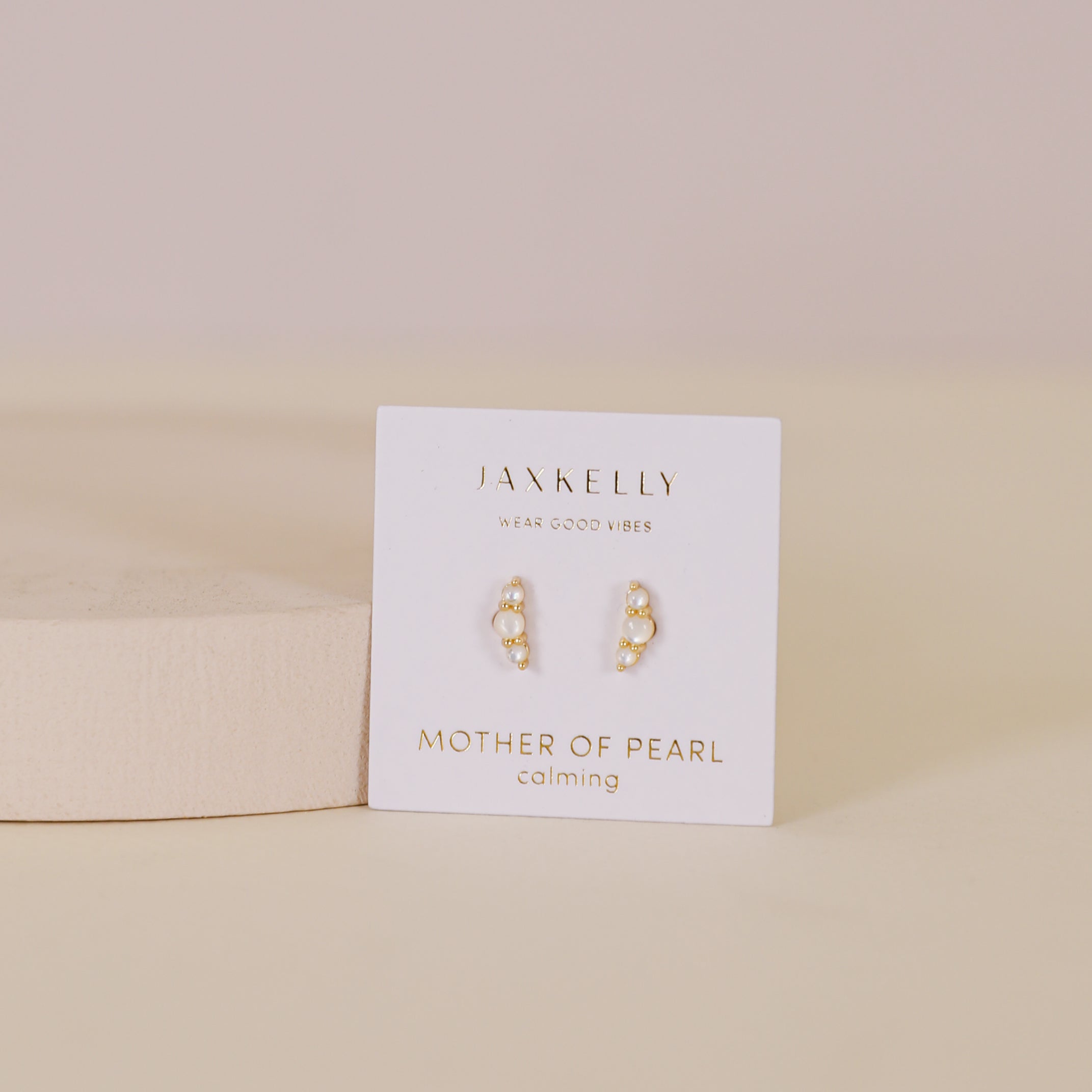A pair of Crescent Gold Earrings - Pearl rests on a white JaxKelly card, displayed on a beige surface with a light pink background.