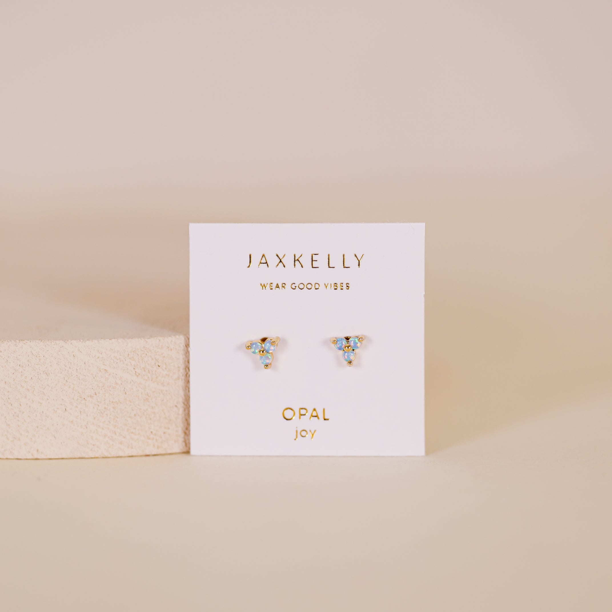 The Trio Cluster Gold Earrings - Fire Opal are shown on a white JaxKelly card marked OPAL joy, set against a soft pink background with a beige textured block on the left.