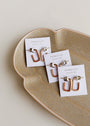 Three pairs of Brown Enamel Large Rectangle Gold Hoop Earrings on JaxKelly cards are displayed on a beige, wavy-edged ceramic dish against a light background.