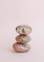 The Moonstone Palm Stone - Luck features three smooth, polished stones in brown and beige hues, including a genuine crystal, stacked against a soft light pink background.