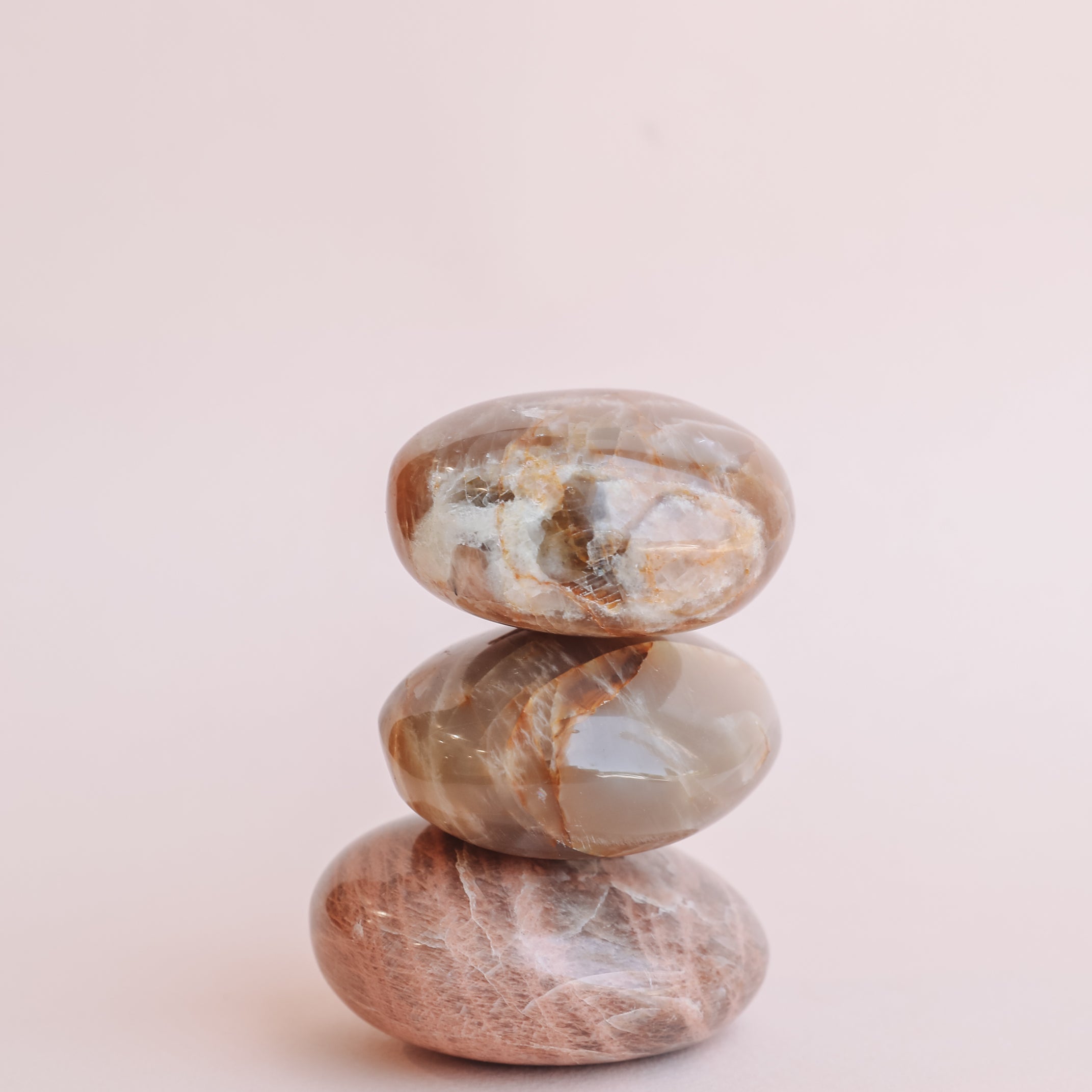 The Moonstone Palm Stone - Luck features three smooth, polished stones in brown and beige hues, including a genuine crystal, stacked against a soft light pink background.