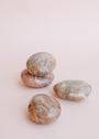 Four Moonstone Palm Stones—three gray with brown and white streaks, one light brown—are arranged on a pink surface. Ideal for manifestation, these genuine crystals for luck feature two stacked and two lying separately.