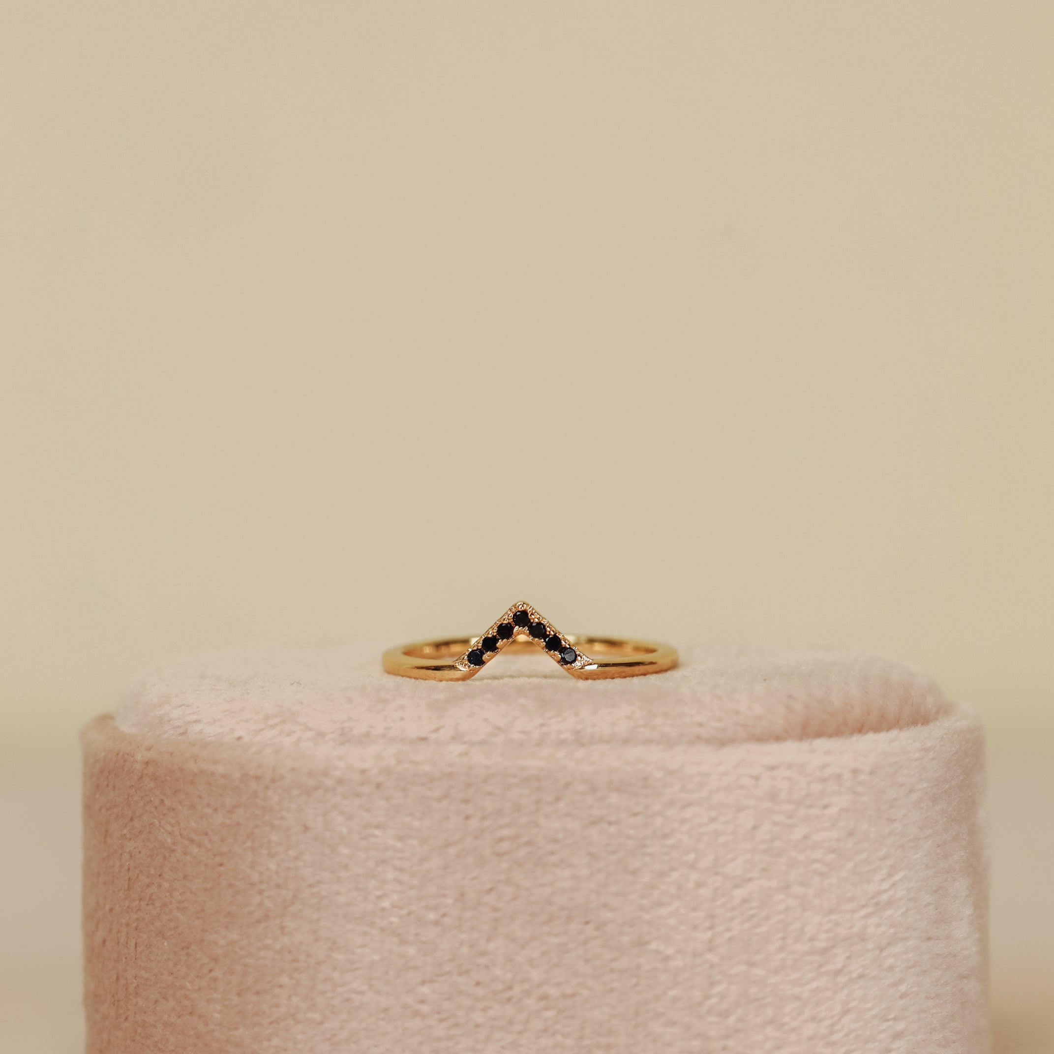 The Black Lambda Ring, a gold plated piece with a chevron design and small dark stones, is shown upright on a soft, round, light pink velvet stand—ideal for stacking against a neutral backdrop.