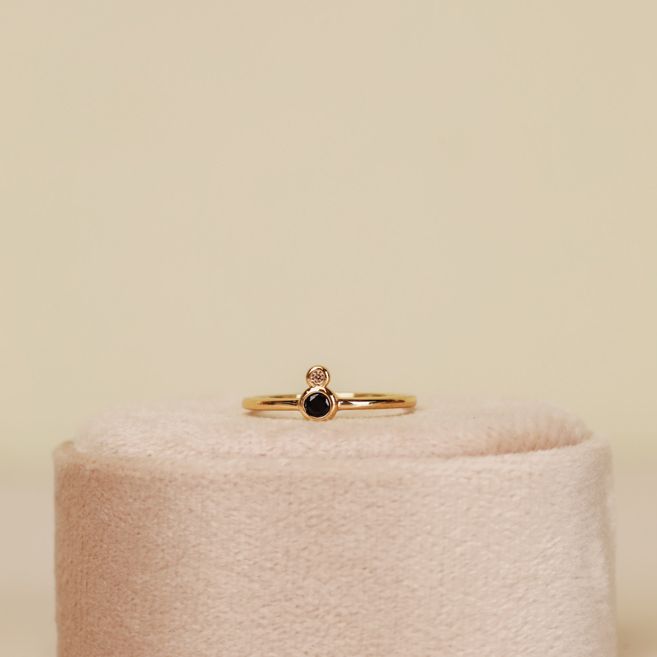 The Black Infinity Ring, an 18k gold plated piece with a small black cubic zirconia and a tiny clear stone above it, is shown upright on a soft pale pink oval ring holder against a neutral background.