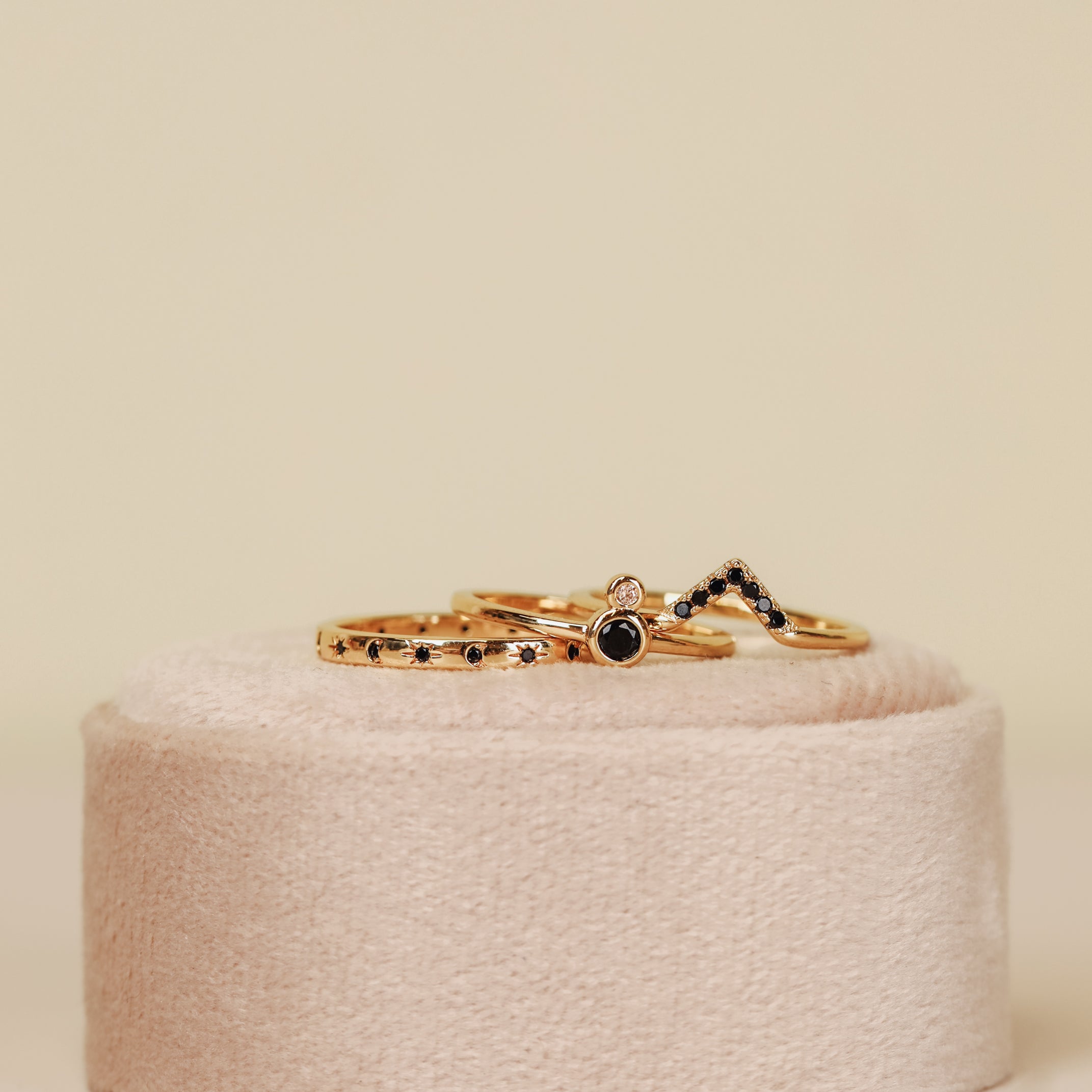 The Black Lambda Ring is a delicate gold plated ring featuring a small black stone, displayed on a soft beige velvet stand, perfect for stacking and adding elegance to any occasion.