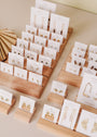 Gold earrings and necklaces, including the Mystery Earring, are displayed on white cards arranged on tiered wooden stands, elegantly presenting a range of jewelry styles in an organized manner.