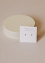 A pair of Teardrop Stud - Clear CZ earrings with 18k gold plating and sterling silver posts are displayed on a white JaxKelly Wear Good Vibes card atop a round, off-white platform on a neutral surface.