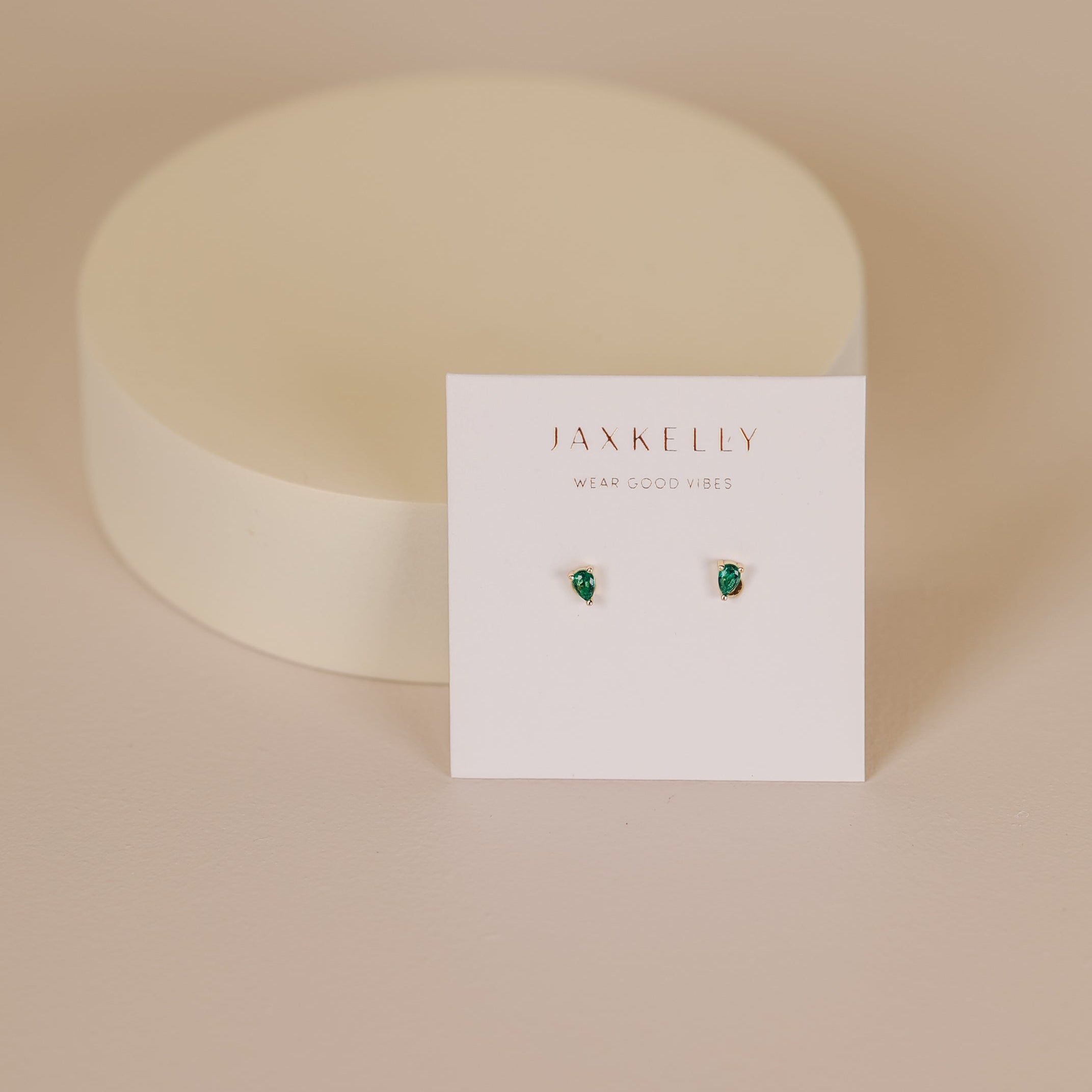 The Teardrop Stud - Emerald CZ earrings are shown on a white JAX KELLY WEAR GOOD VIBES card, set against a soft beige background with a round, off-white prop behind them.