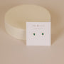 The Teardrop Stud - Emerald CZ earrings are shown on a white JAX KELLY WEAR GOOD VIBES card, set against a soft beige background with a round, off-white prop behind them.
