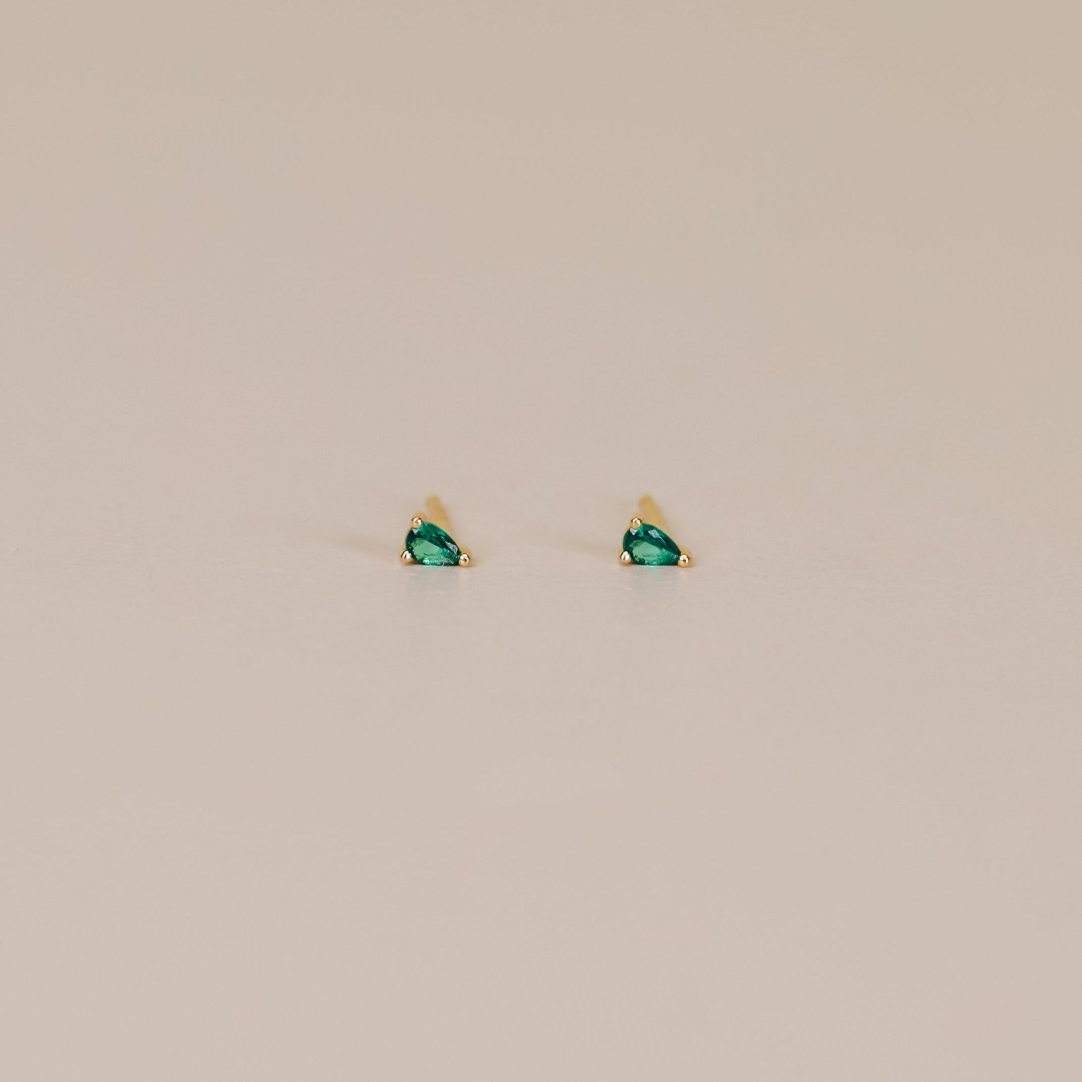 The Teardrop Stud - Emerald CZ earrings feature small 18k gold plating and sparkling green CZ stones, shown against a plain beige background.