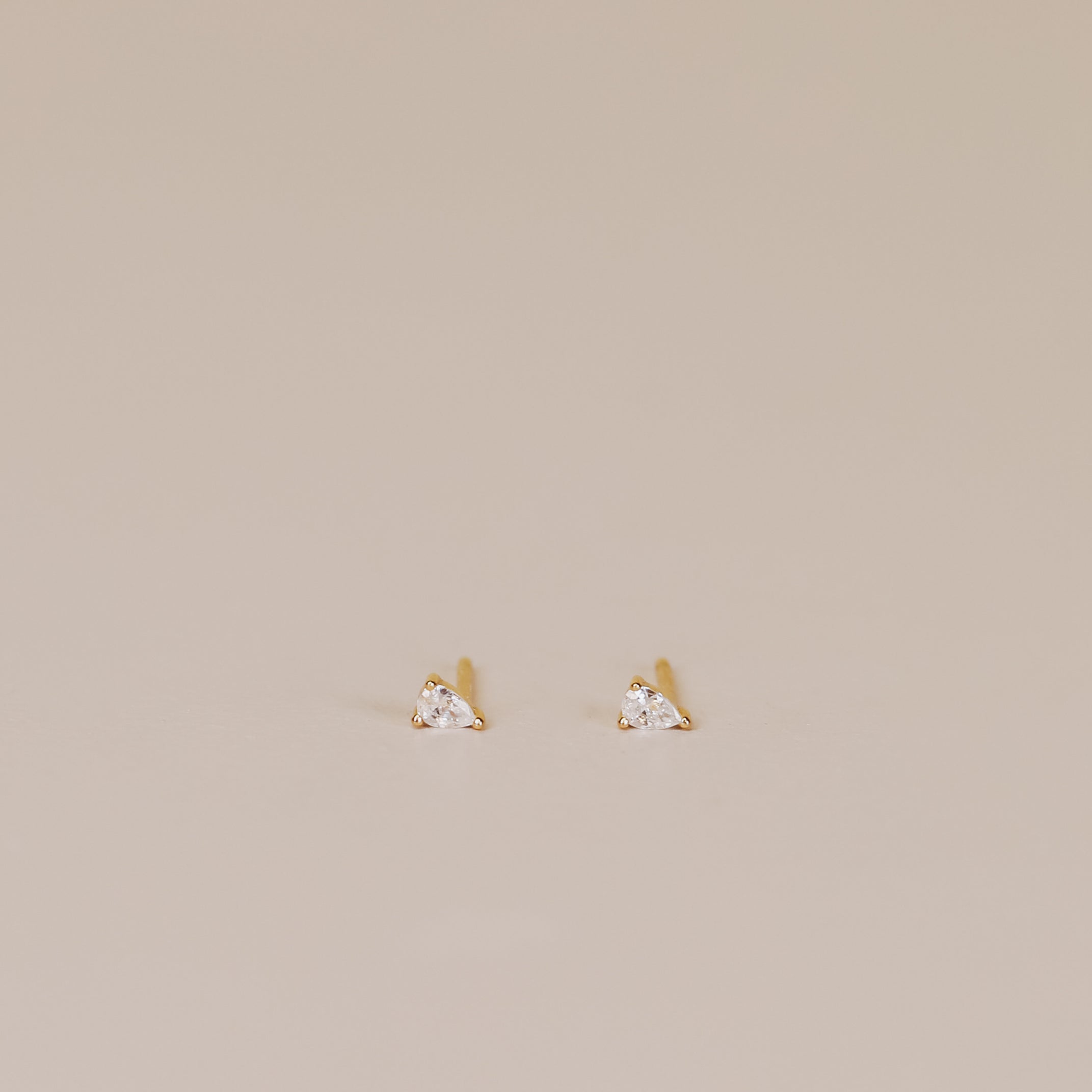 The Teardrop Stud - Clear CZ features small, gold-plated stud earrings with clear cubic zirconia and sterling silver posts, shown on a light beige background.