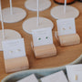 Three pairs of stud earrings on white "JaxKelly" cards in wooden stands; one pair is the Teardrop Stud - Emerald CZ, featuring green teardrop stones, while the others are simple metallic studs with 18k gold plating.