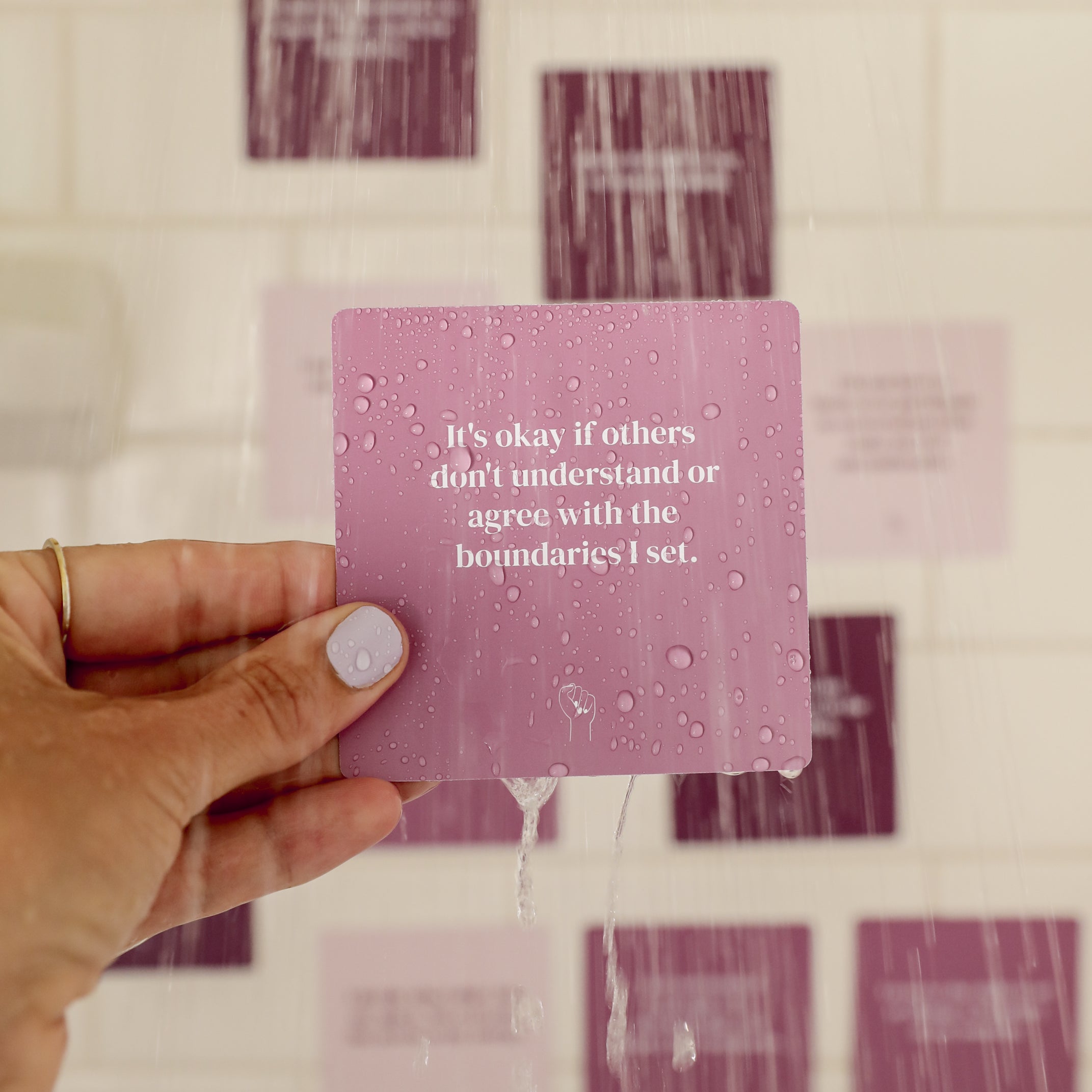 A hand with painted nails holds a wet purple Boundaries Shower Affirmations™ card in a shower. The card reads, "It's okay if others don't understand or agree with the boundaries I set." Blurry cards are visible on white tiles behind.