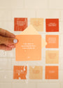 A hand with light purple nails holds a peach Positivity Shower Affirmations™ waterproof card reading “I am open to receiving the love I am worthy of,” with blurred affirmation cards on a white tiled wall in the background.