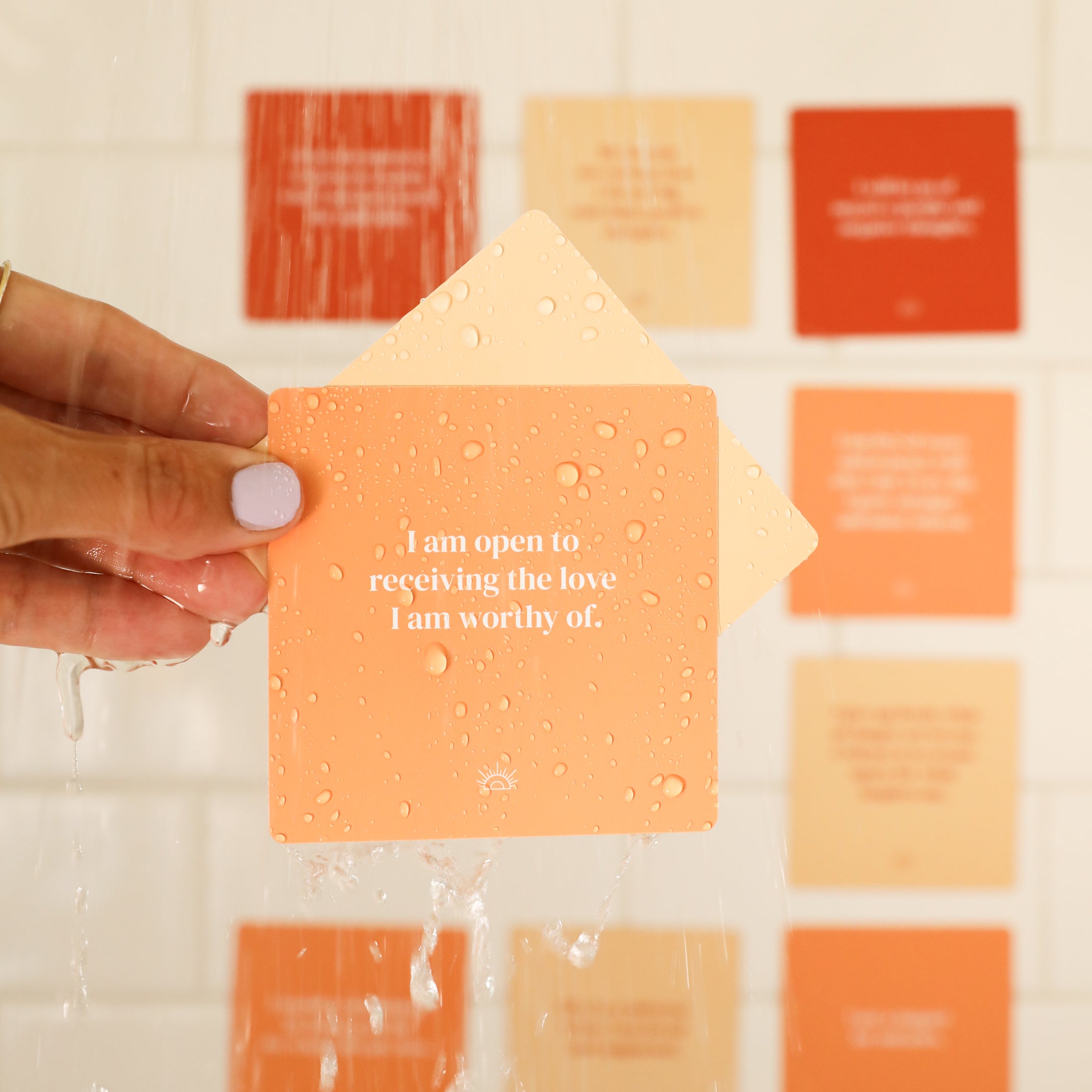 A hand with light purple nails holds a peach Positivity Shower Affirmations™ waterproof card reading “I am open to receiving the love I am worthy of,” with blurred affirmation cards on a white tiled wall in the background.