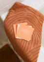 Three Positivity Shower Affirmations™ waterproof quote cards with uplifting messages sit on a textured rust towel over a bathtub edge. The top card reads, “I manifest abundance by being grateful for what I already have.”.