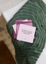 A stack of Boundaries Shower Affirmations™ waterproof cards rests on a green towel in a bathroom with white tiles and a woven rug. The top card reads, "I no longer say yes so that others will feel comfortable.