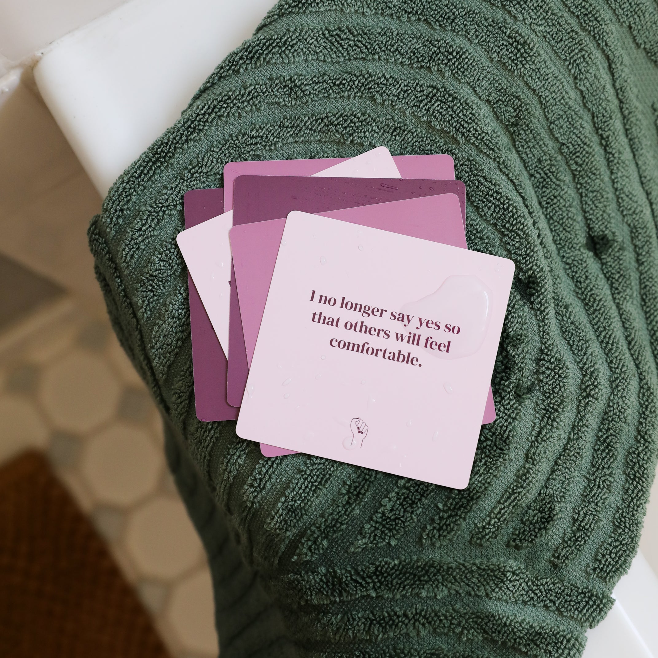 A stack of Boundaries Shower Affirmations™ waterproof cards rests on a green towel in a bathroom with white tiles and a woven rug. The top card reads, "I no longer say yes so that others will feel comfortable.