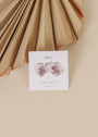 A pair of gold-plated Amethyst Gemstone Prong - Healing star-shaped stud earrings are displayed on a white card with "wear good vibes" and "AMETHYST healing" text, set against a neutral background with pleated decor.