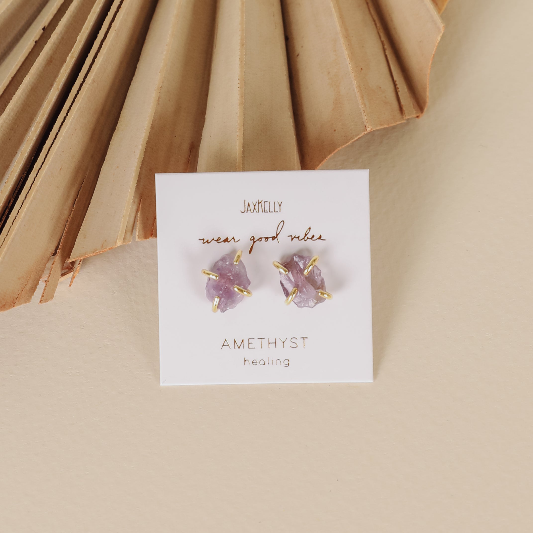 A pair of gold-plated Amethyst Gemstone Prong - Healing star-shaped stud earrings are displayed on a white card with "wear good vibes" and "AMETHYST healing" text, set against a neutral background with pleated decor.
