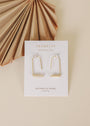 A pair of Mother of Pearl Gemstone Drop - Calming earrings with gold plating, displayed on a white card labeled JaxKelly and Mother of Pearl, set on a beige background beside a dried palm leaf.