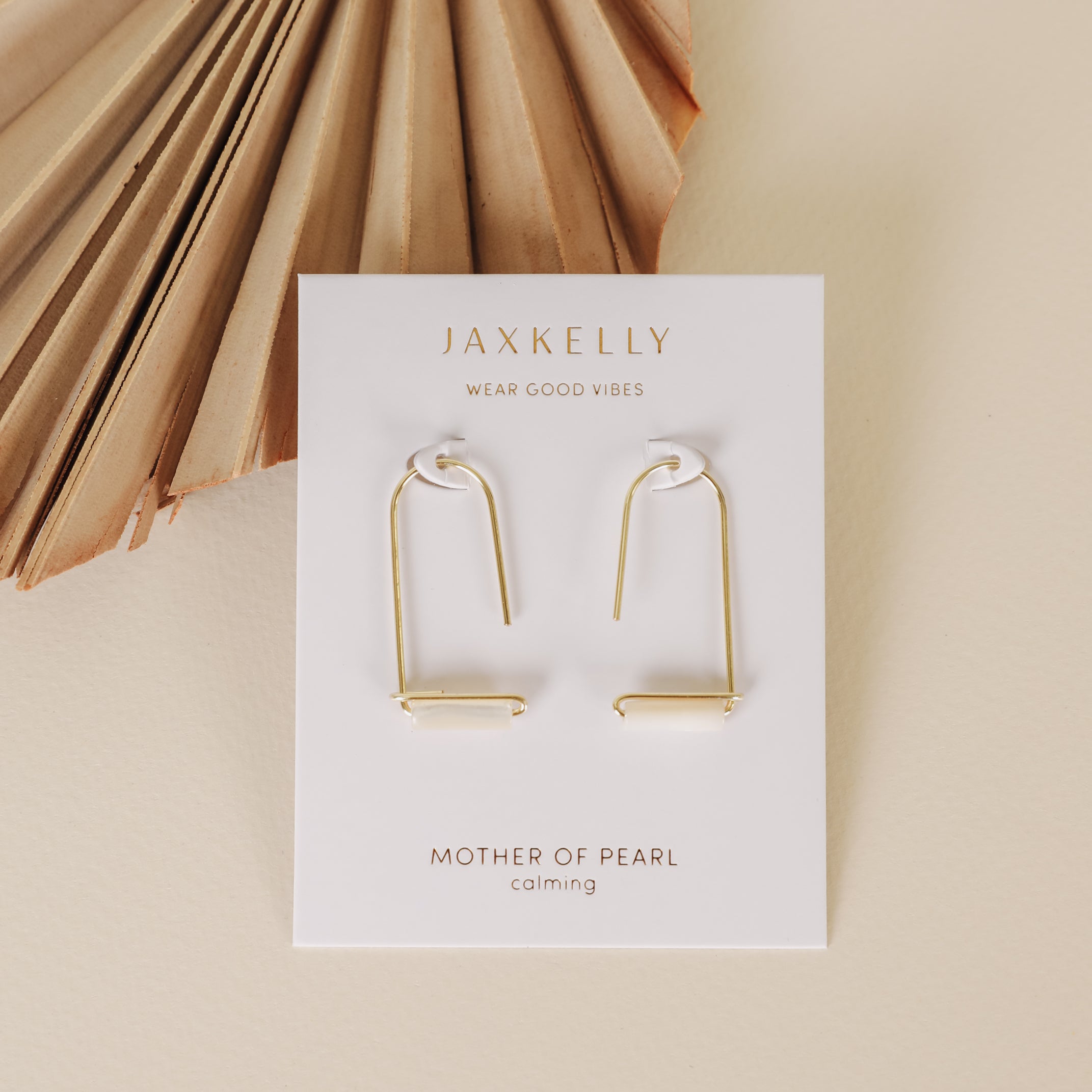 A pair of Mother of Pearl Gemstone Drop - Calming earrings with gold plating, displayed on a white card labeled JaxKelly and Mother of Pearl, set on a beige background beside a dried palm leaf.