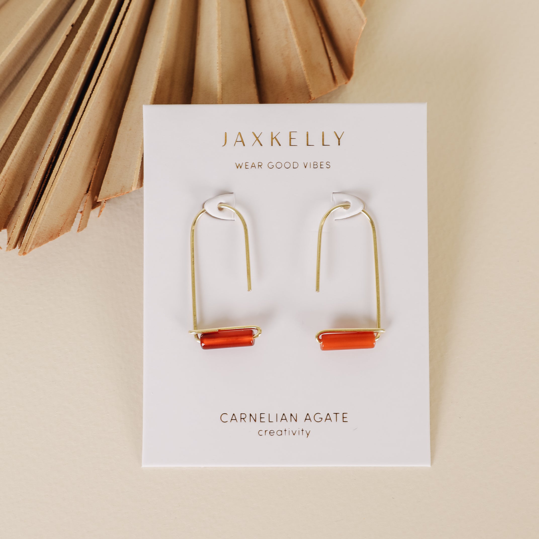 Carnelian Agate Gemstone Drop - Creativity earrings featuring rectangular orange stones on a JaxKelly card, displayed on a beige surface with a dried palm leaf in the corner.