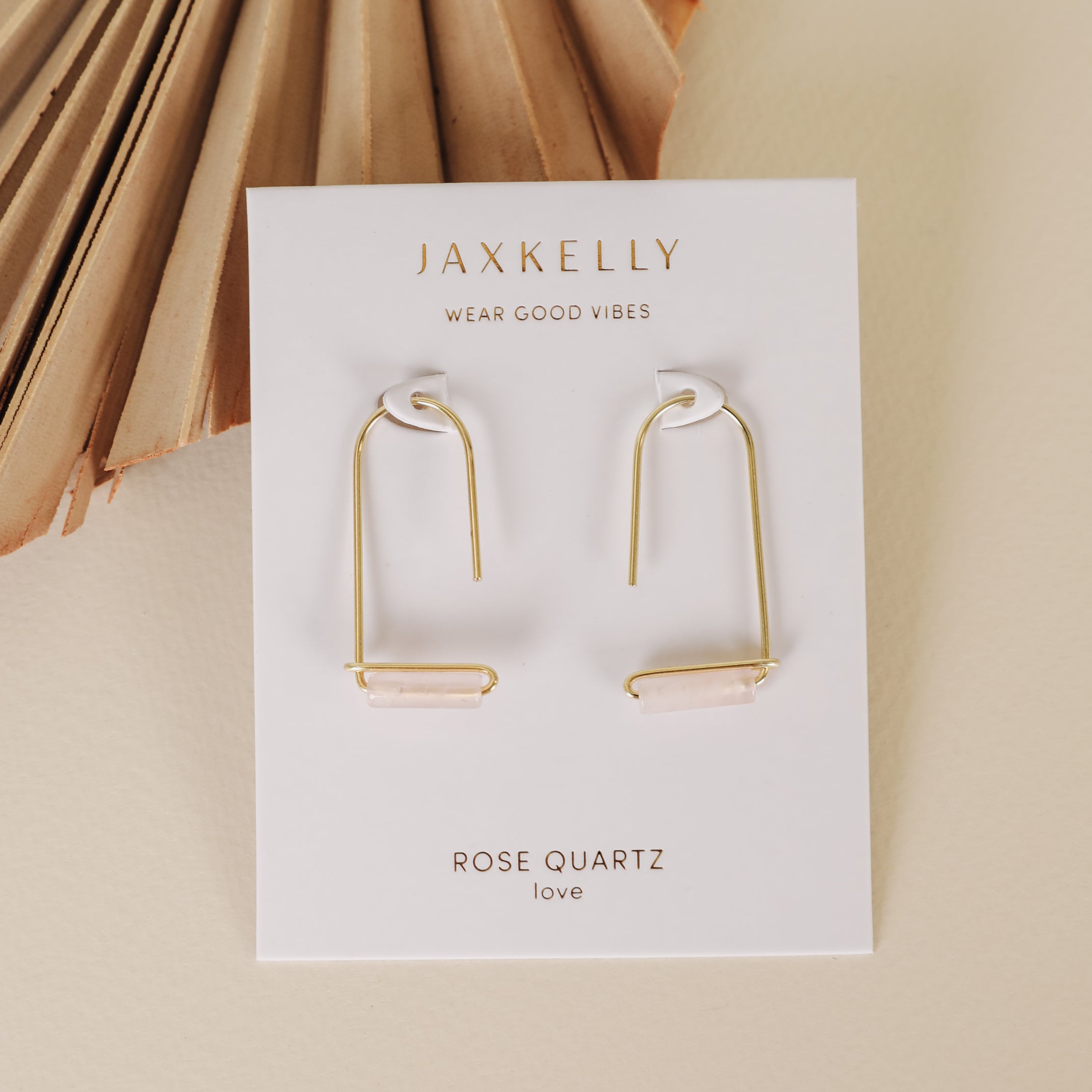 The Rose Quartz Gemstone Drop - Love earrings feature 18k gold plating with horizontal rose quartz bars, displayed on a JaxKelly card reading “Wear Good Vibes and Rose Quartz love,” set on a beige surface with a paper fan decoration.