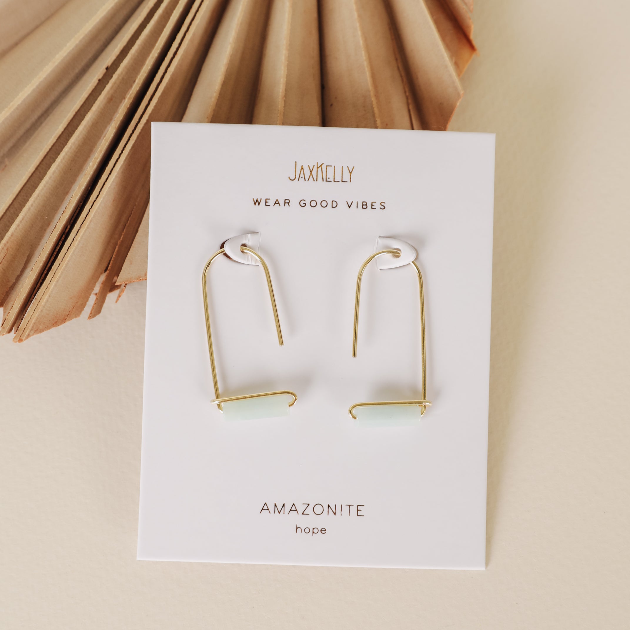 A pair of elegant 18k gold plated Amazonite Gemstone Drop - Hope earrings with rectangular stones is displayed on a white JaxKelly card, placed beside a folded brown decorative fan.
