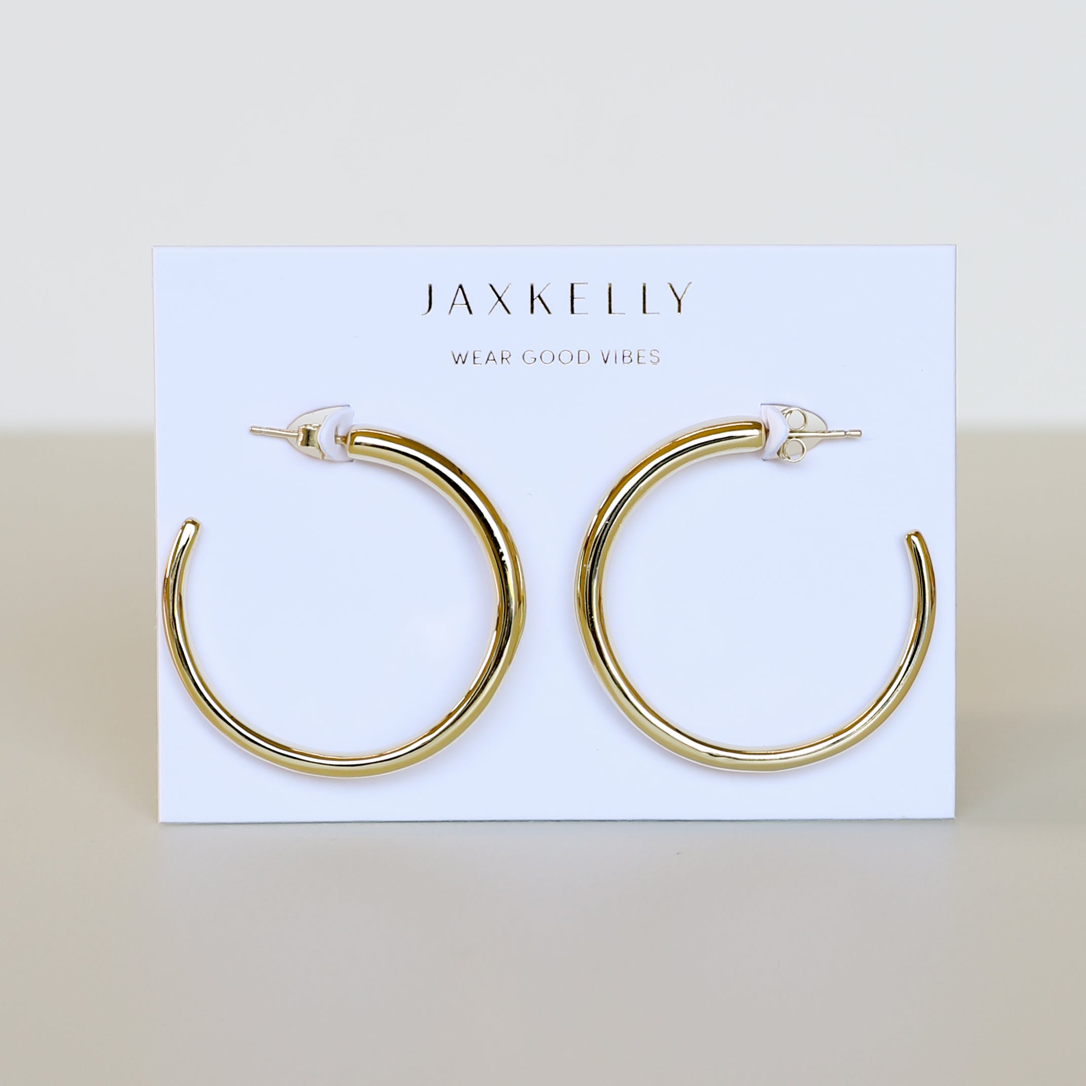 The Gold Everyday Hoop - Large, 18k gold plated bold hoops, are displayed on a white card with JaxKelly and Wear Good Vibes printed on it against a neutral background.
