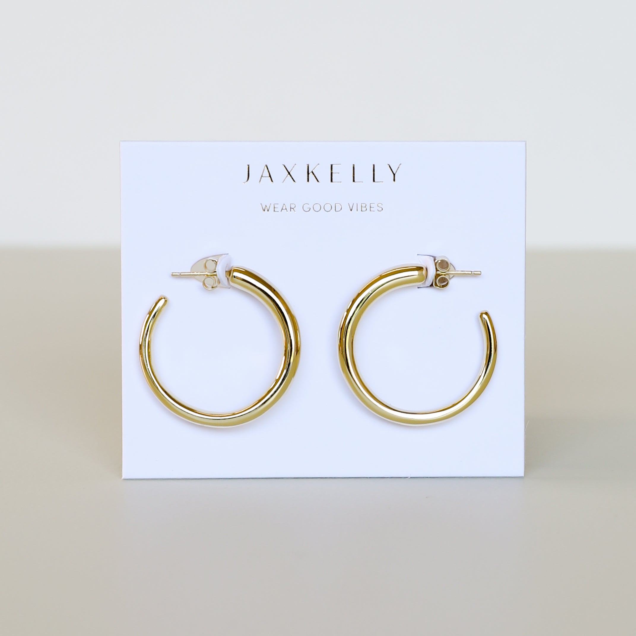 A pair of Gold Everyday Hoop - Medium earrings displayed on a white card featuring the JAX KELLY brand and Wear Good Vibes slogan, highlighting these timeless jewelry essentials.