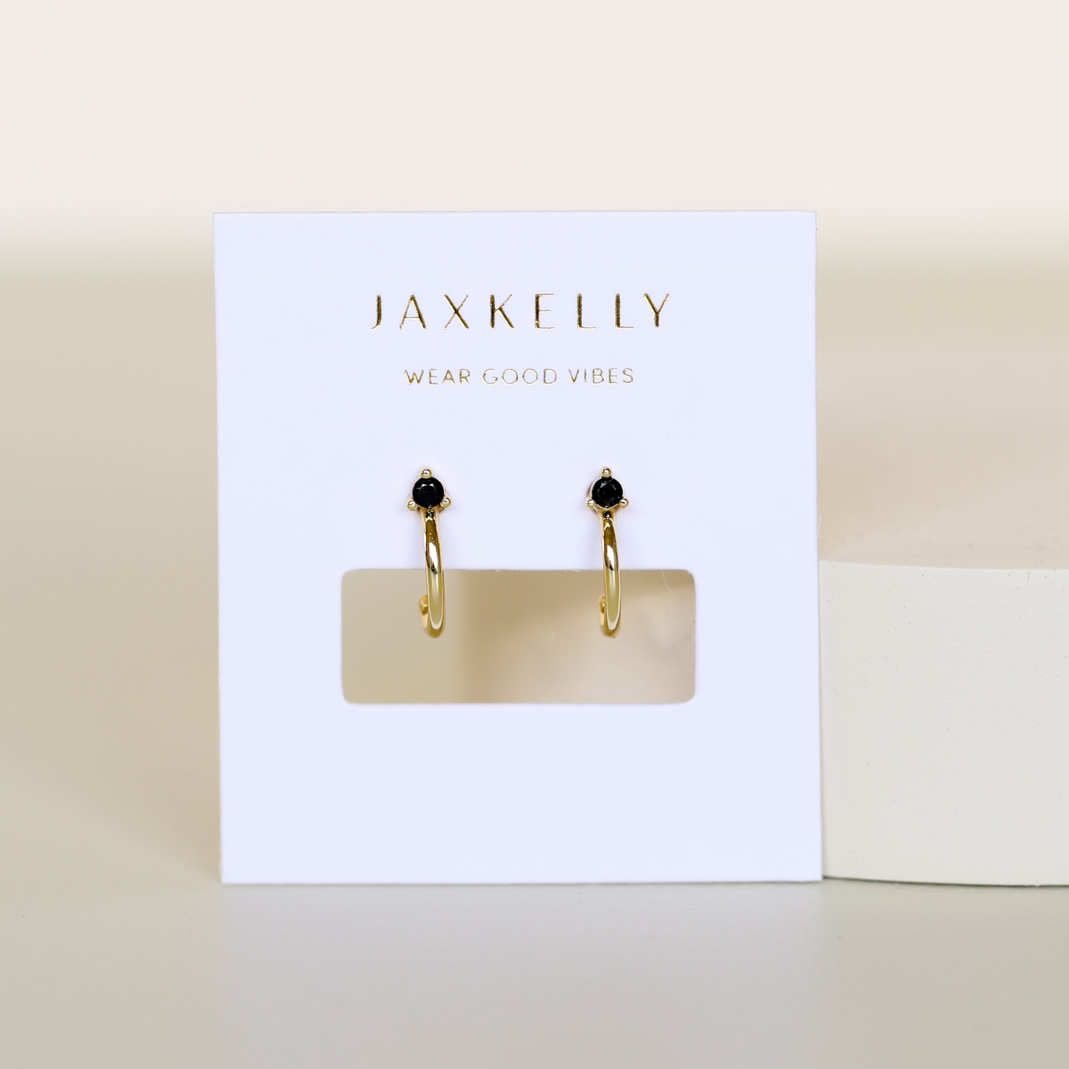 The Open Mini Hoop Earring - Black features gold-plated hoops with small black CZ stones, displayed on a white JaxKelly card that says WEAR GOOD VIBES in gold, set against a minimal cream background.