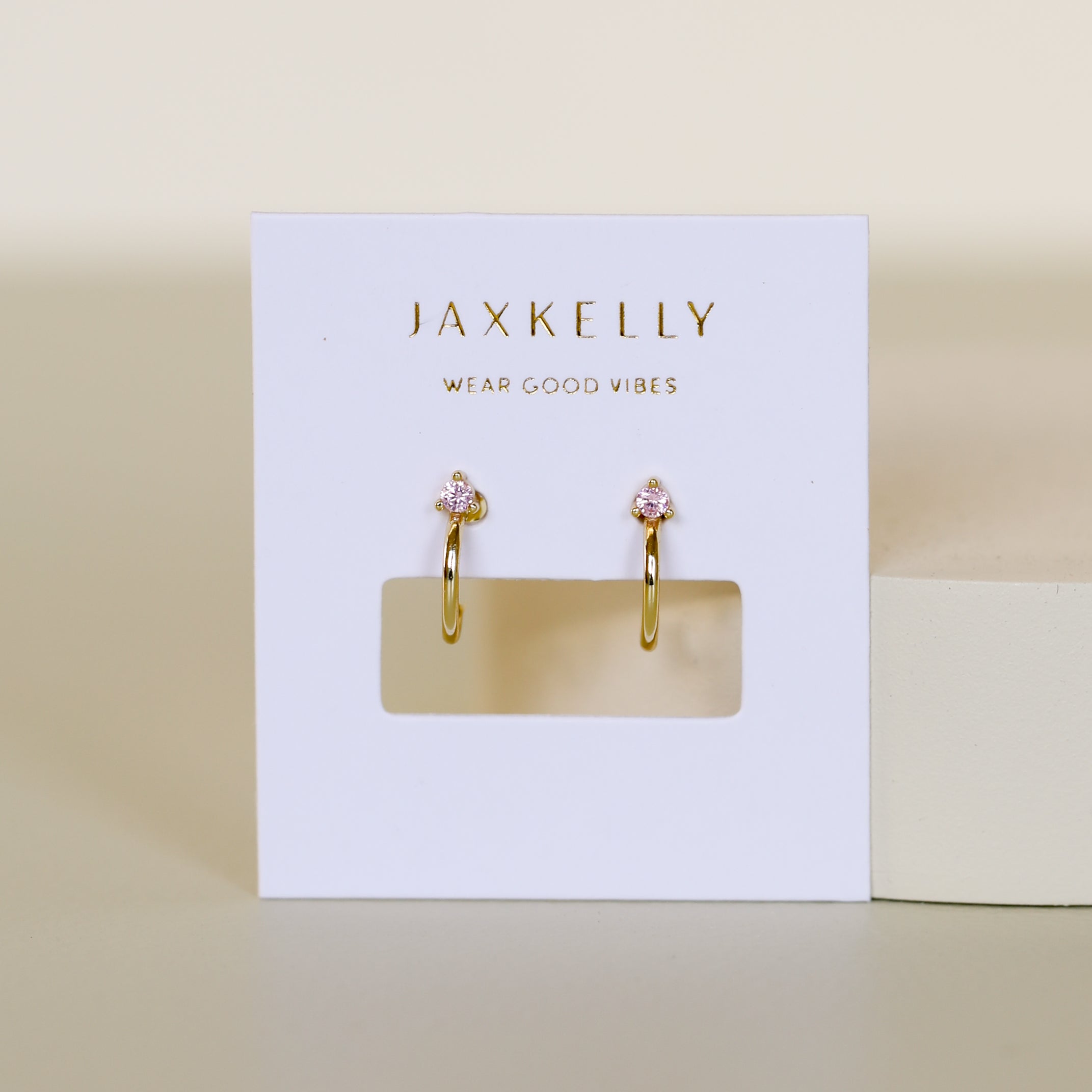 The Open Mini Hoop Earring - Pink features gold hoops adorned with small pink CZ stones, displayed on a white JaxKelly card reading "WEAR GOOD VIBES" in gold lettering. The neutral background enhances its minimalistic look.