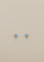 The Simple Stud Earring - Fire Opal features round blue lab-grown fire opal gemstones set in the center of 18k gold plated studs, shown on a plain beige background.