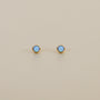 The Simple Stud Earring - Fire Opal features round blue lab-grown fire opal gemstones set in the center of 18k gold plated studs, shown on a plain beige background.