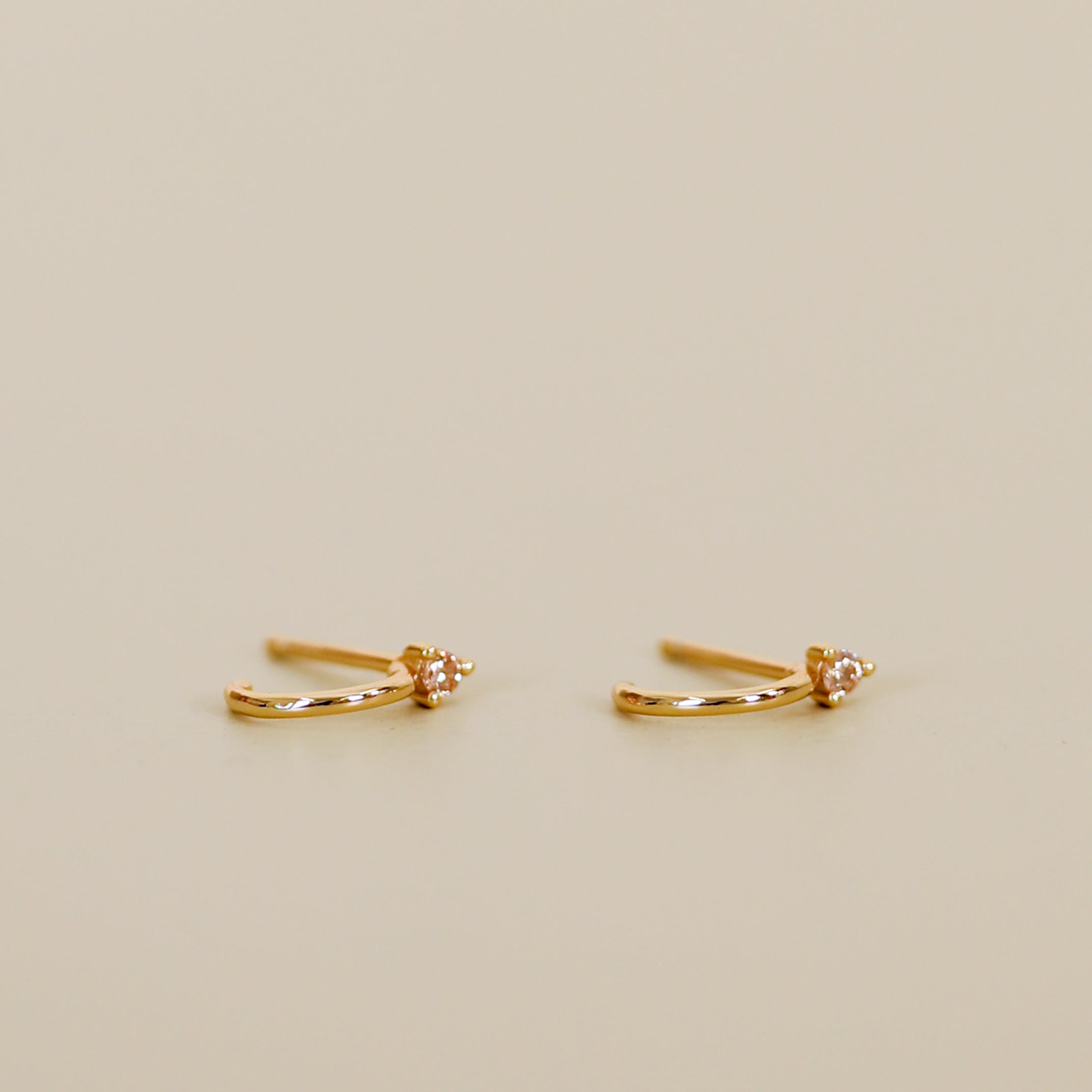 The Open Mini Hoop Earring - Champagne features delicate 18K gold plating and a round champagne CZ stone, styled as small hoops and showcased on a plain beige background.