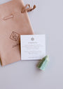 A Mini Amazonite Quartz Crystal Point, a suede pouch, and an info card with minimalist text and graphics on amazonite's calming properties rest on a light surface.