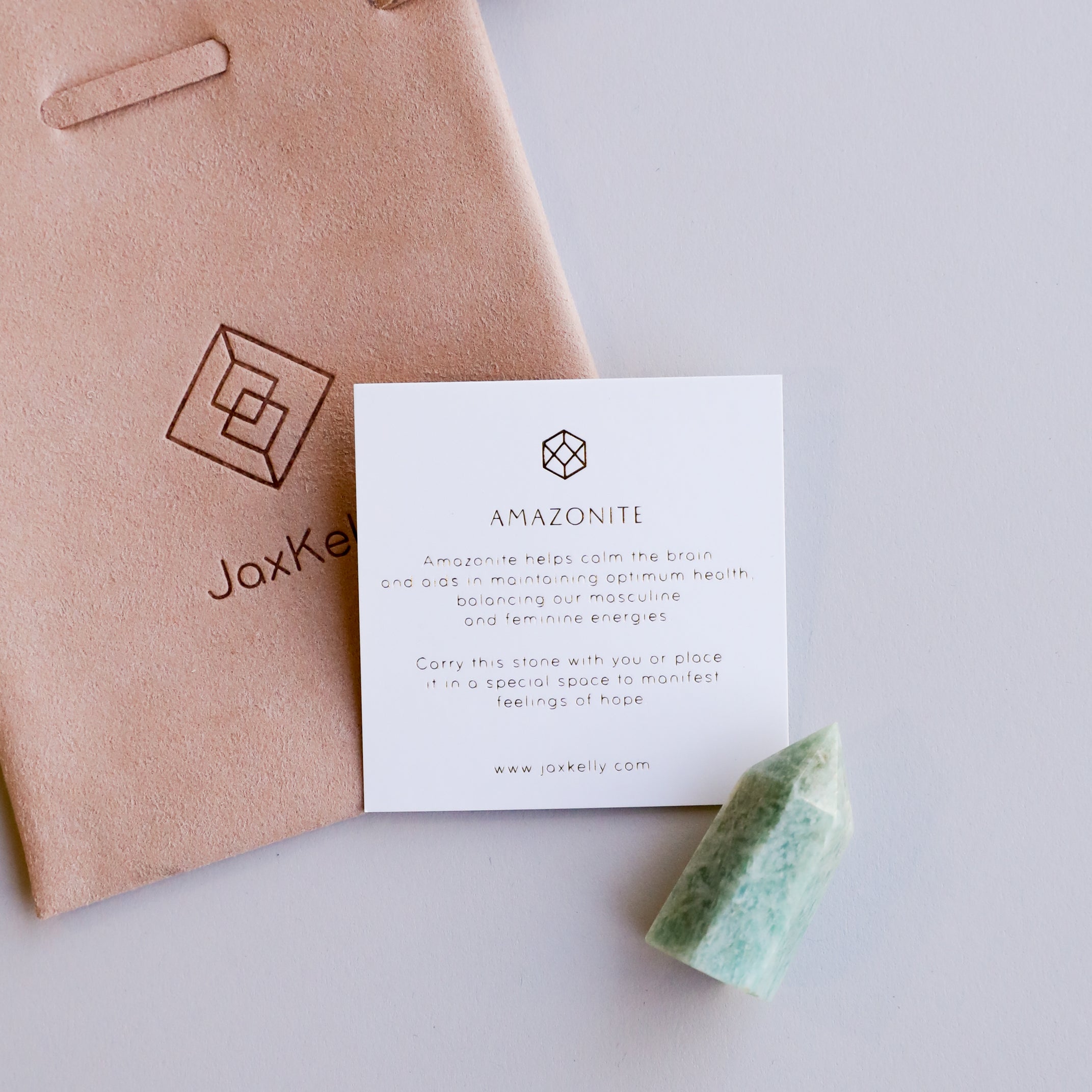 A Mini Amazonite Quartz Crystal Point, a suede pouch, and an info card with minimalist text and graphics on amazonite's calming properties rest on a light surface.