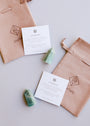 Two green Mini Amazonite Quartz Crystal Points rest on a white surface beside beige “JaxKelly” drawstring pouches and informational cards that detail the calming properties of this healing stone.