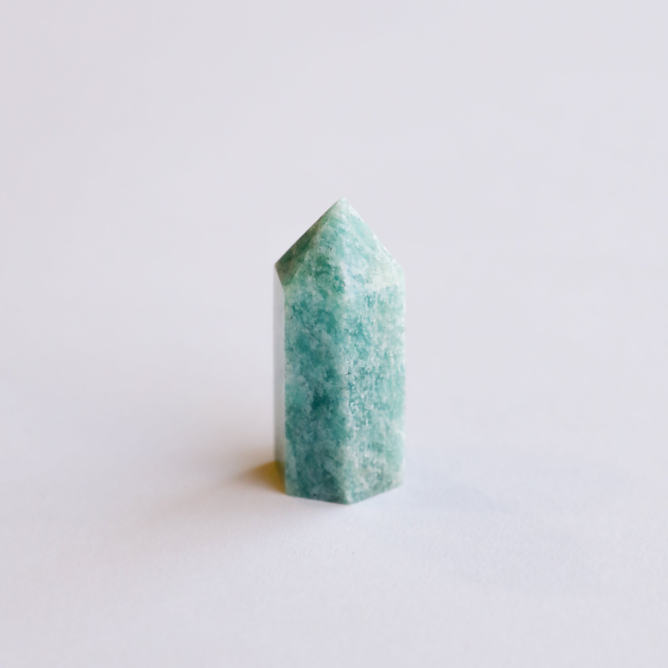 The Mini Amazonite Quartz Crystal Point stands upright on a white surface, displaying its polished finish and soothing blue-green hue, perfect for adding a calming touch to any space.