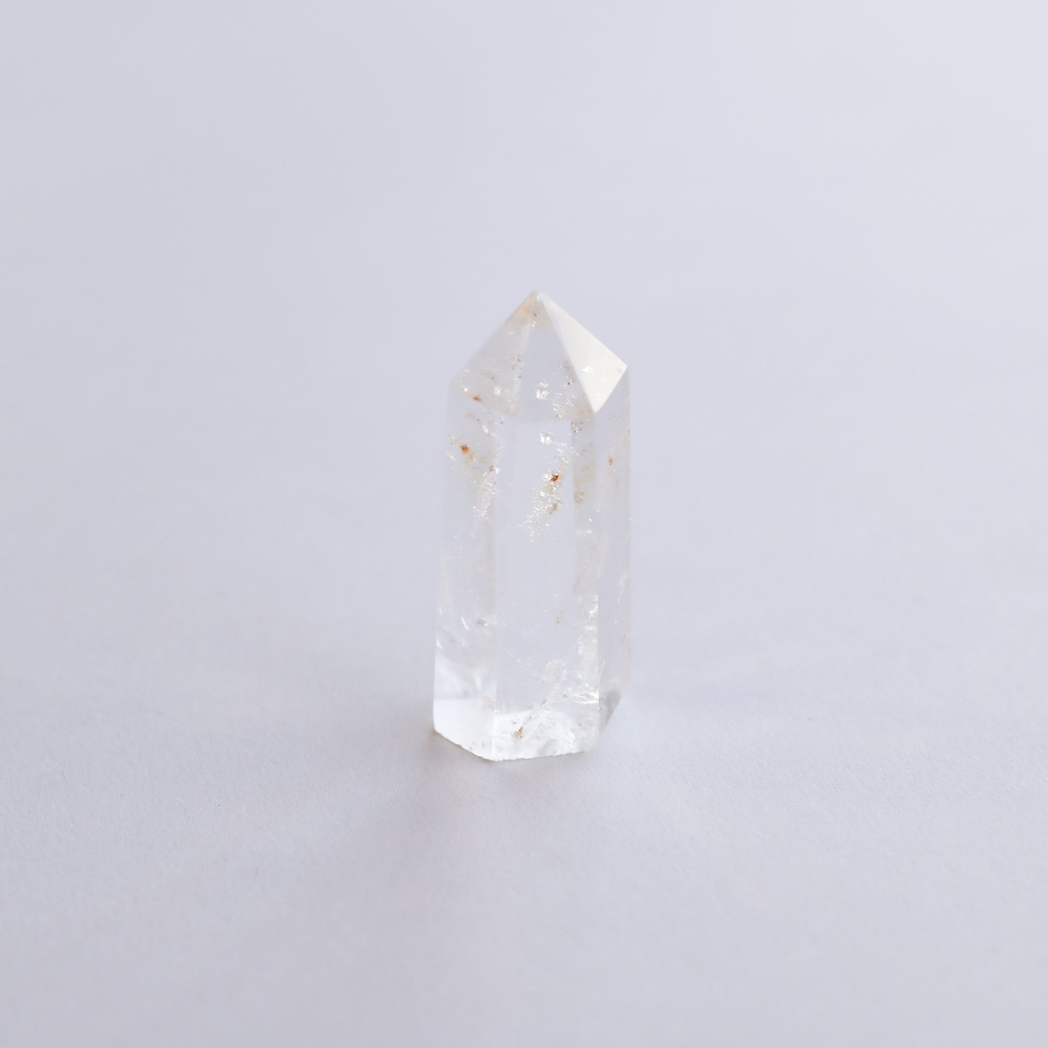 The Mini Clear Quartz Crystal Point, featuring small inclusions, stands upright on a light-colored surface against a soft, minimal background.