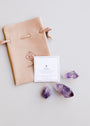 The Raw Amethyst Crystal Set includes a tan suede drawstring pouch with a geometric JaxKelly logo, a white card labeled “AMETHYST,” and three raw amethyst stones—believed to support intuition and healing—displayed on light grey.