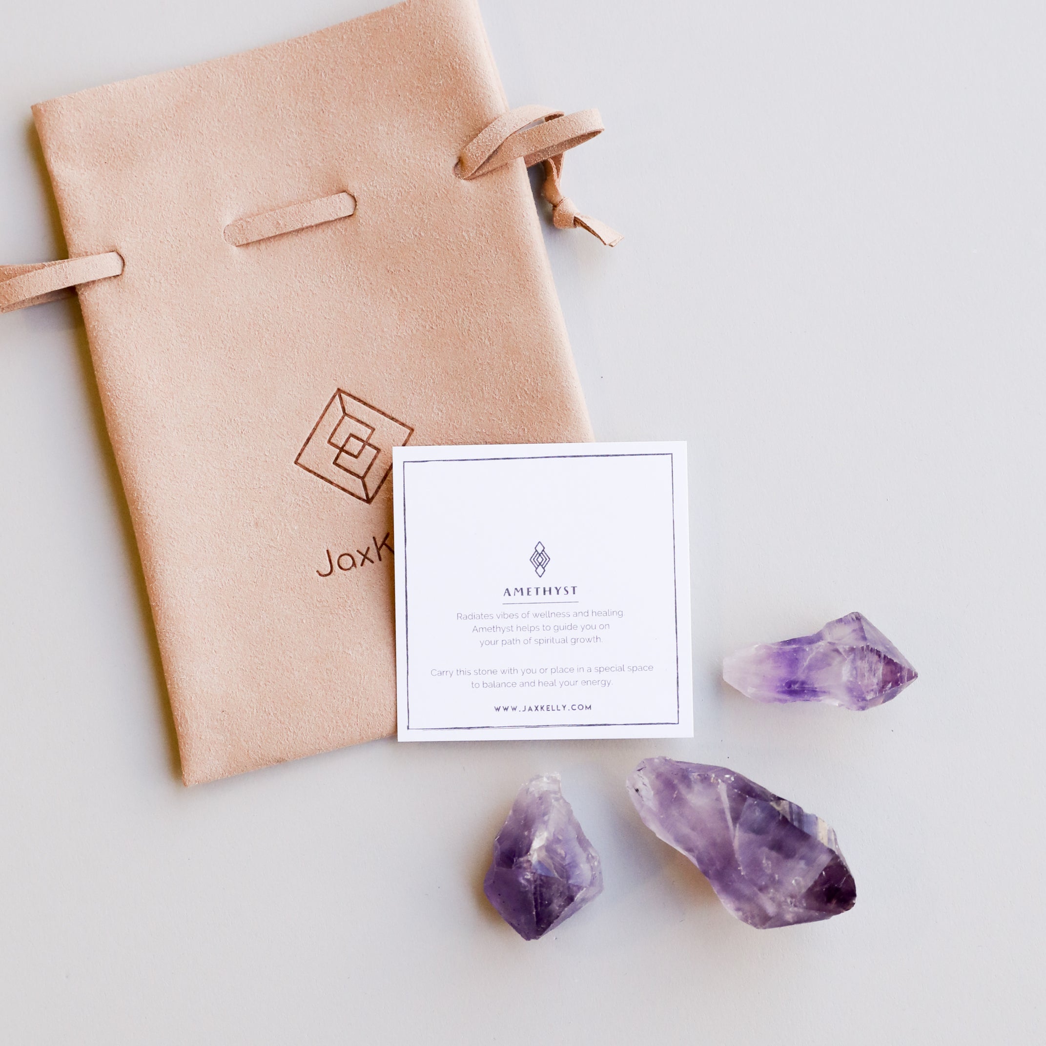 The Raw Amethyst Crystal Set includes a tan suede drawstring pouch with a geometric JaxKelly logo, a white card labeled “AMETHYST,” and three raw amethyst stones—believed to support intuition and healing—displayed on light grey.