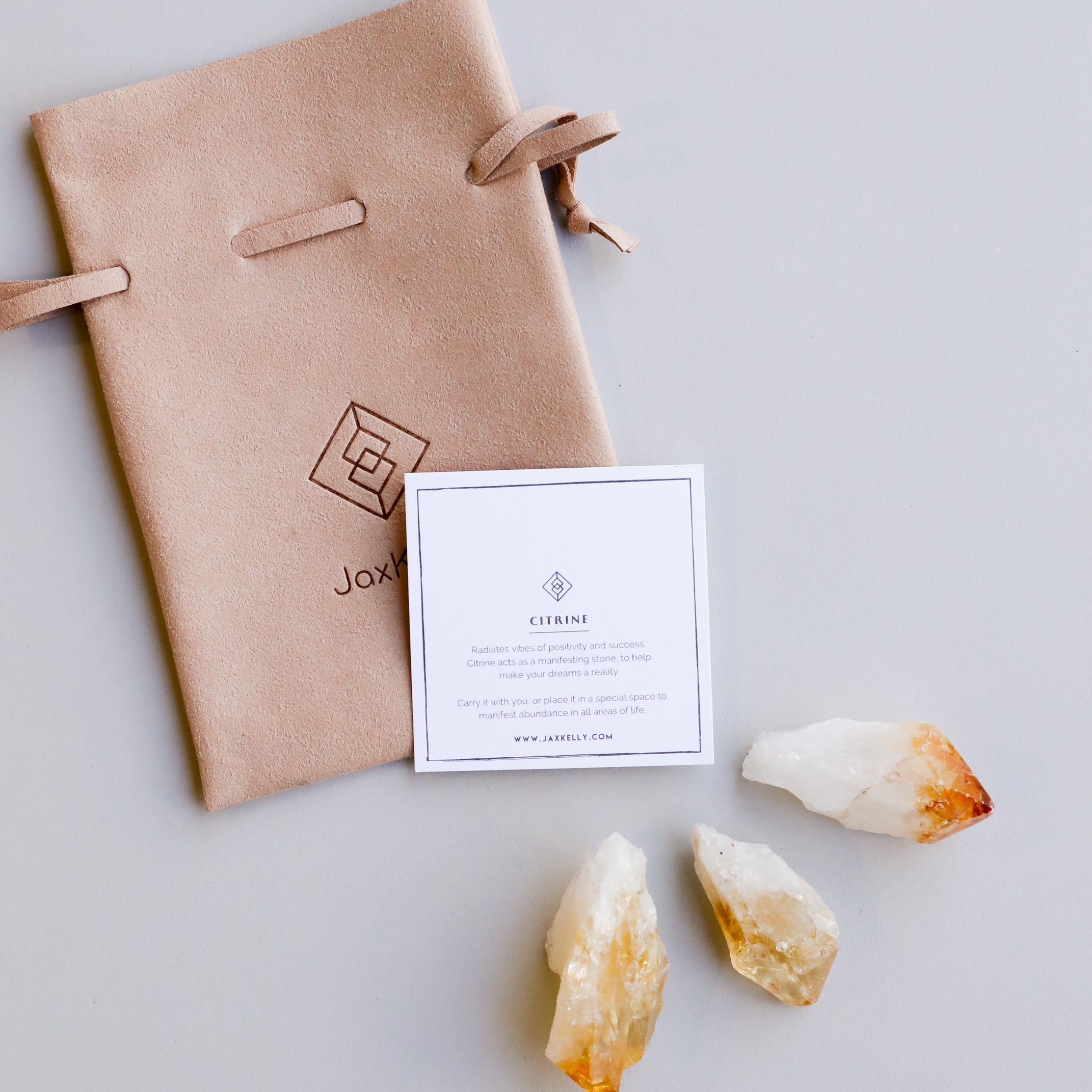 The Raw Citrine Crystal Set includes a beige suede drawstring pouch, an informational card about citrine, and three raw Citrine stones, all displayed on a light gray surface.